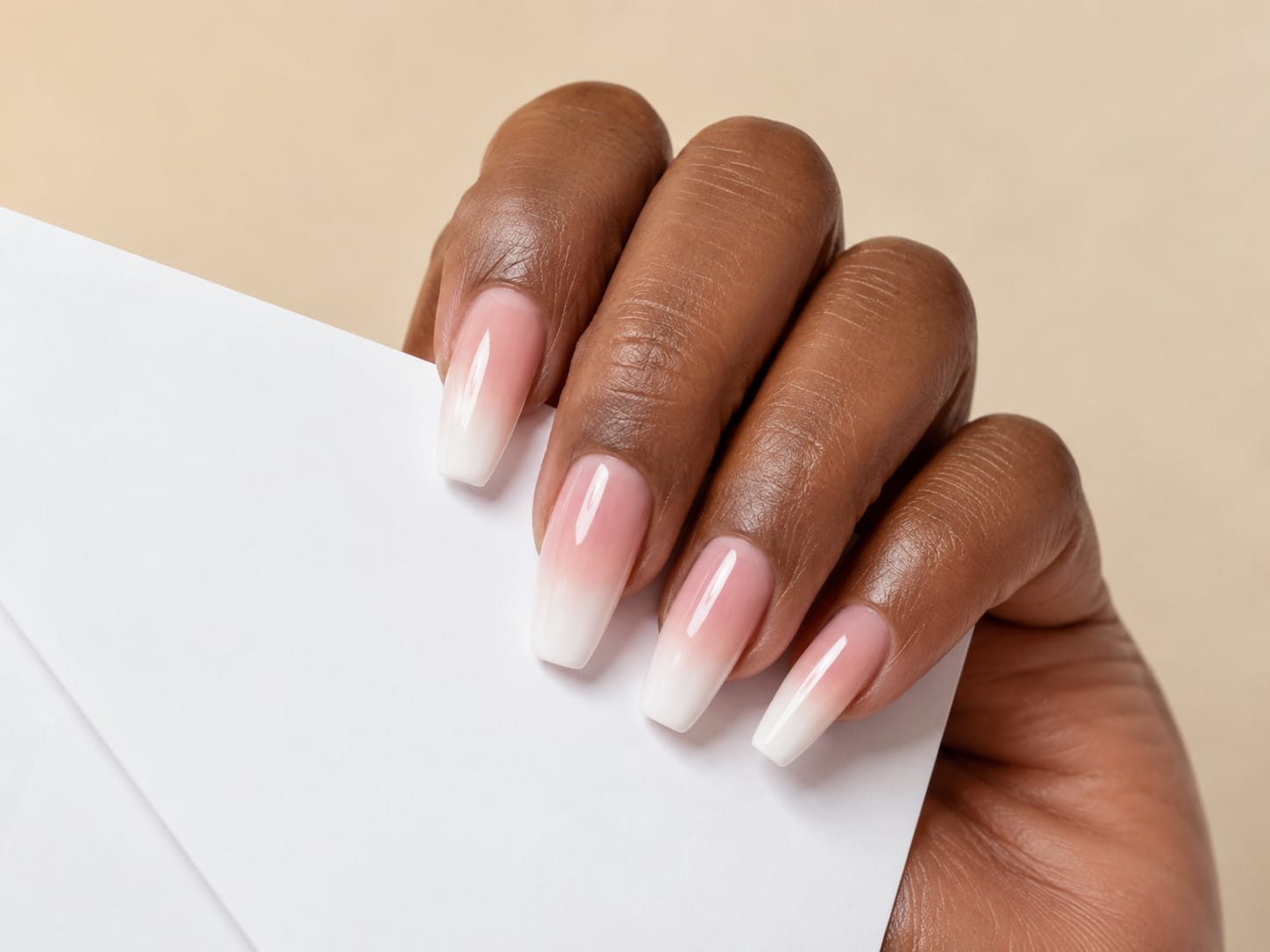 Long coffin pink baby-boomer ombre French nails on a dark-skinned hand holding a white envelope.