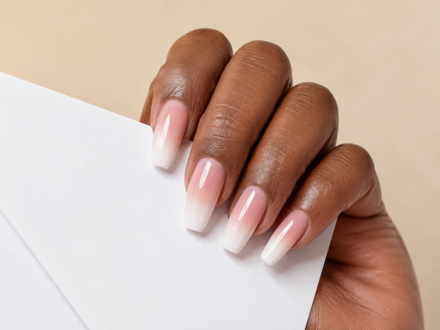 Long coffin pink baby-boomer ombre French nails on a dark-skinned hand holding a white envelope.