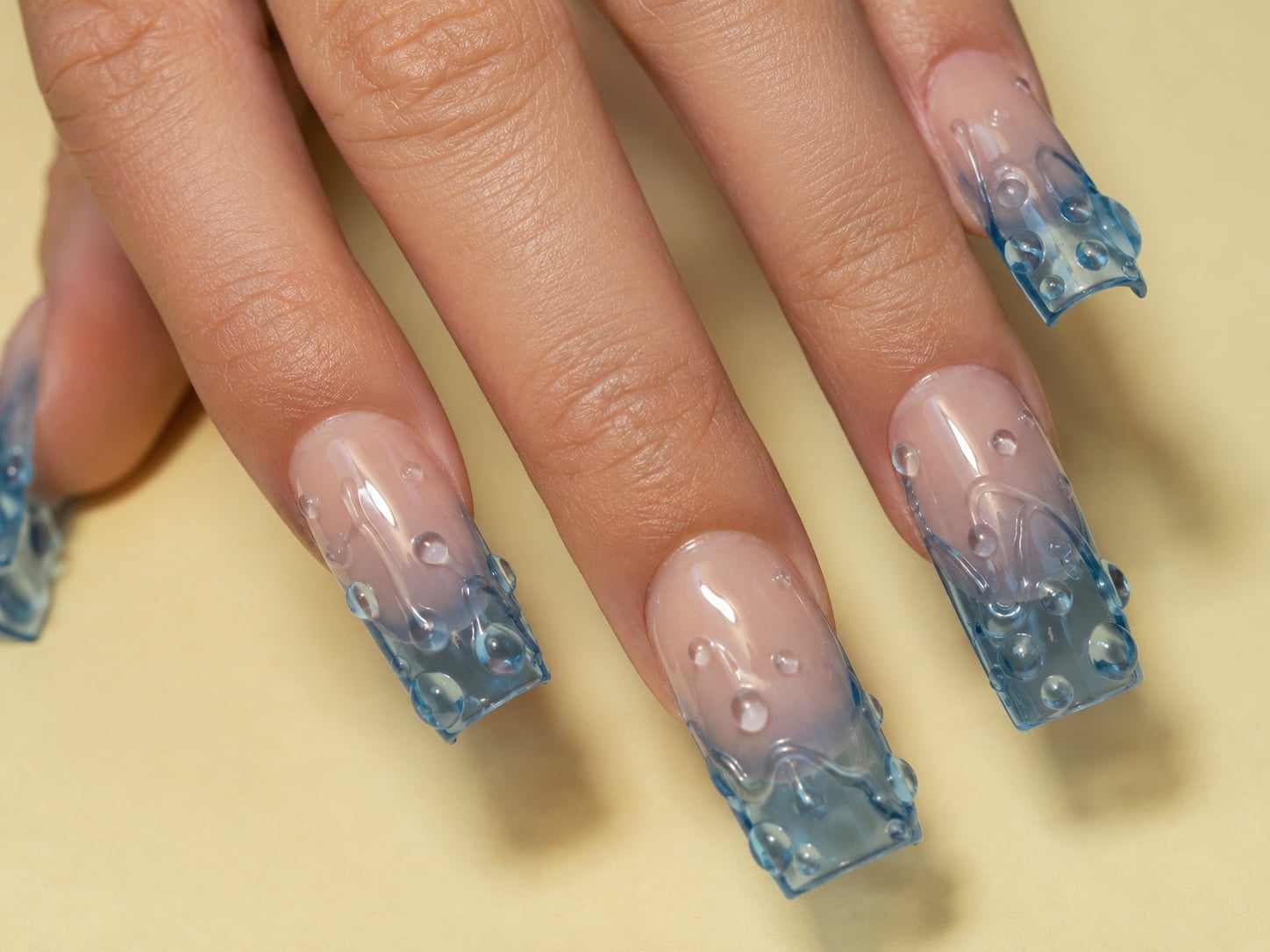 Long coffin sheer nails with 3D blue water-droplet textured tips against a tan background.