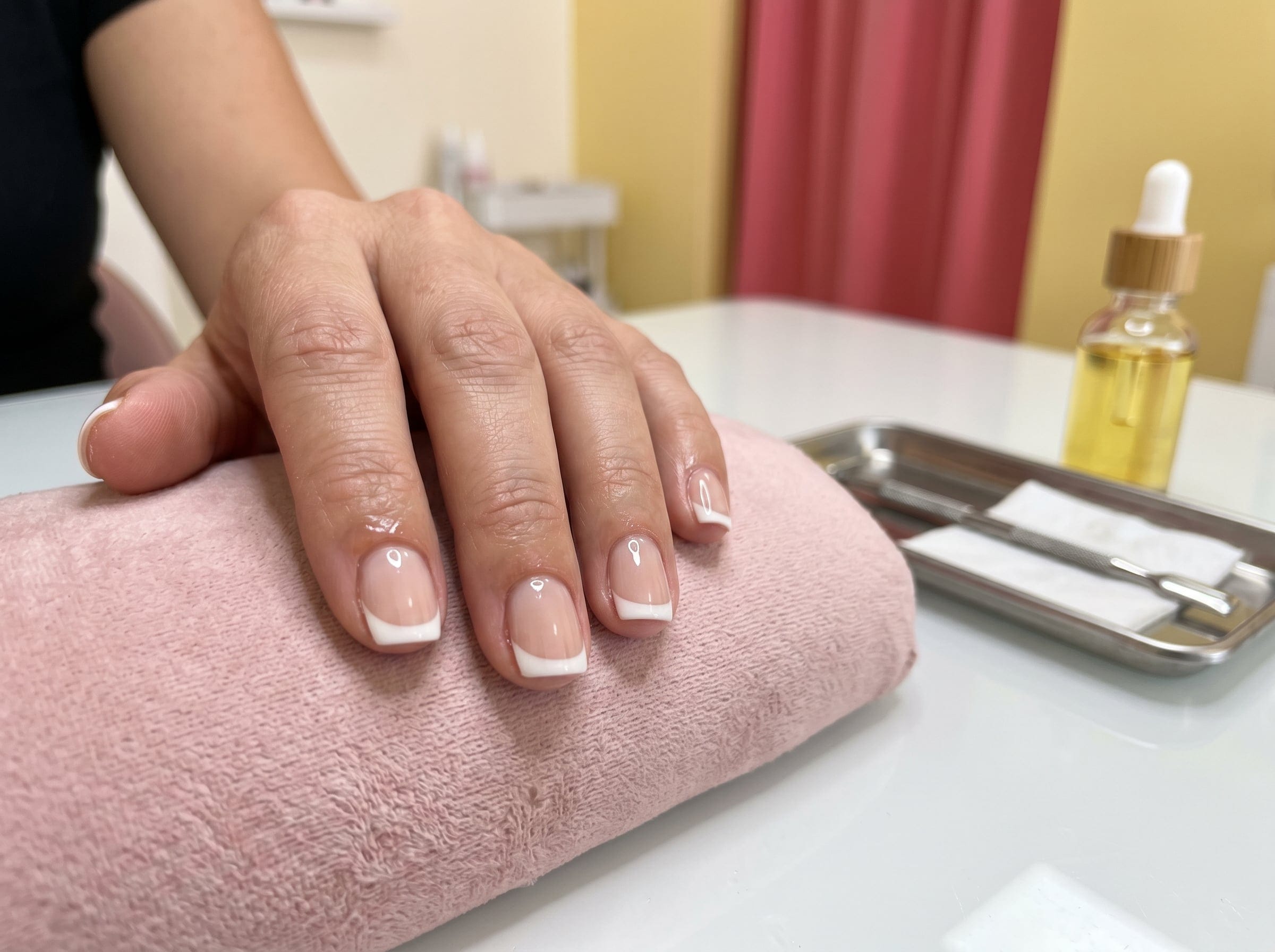 Soft white-tip French manicure on short square nails resting on a pink towel beside a cuticle oil tray.