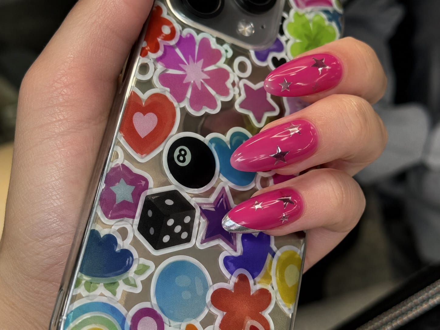 Long almond hot-pink nails with silver star accents holding a sticker-covered phone case selfie.