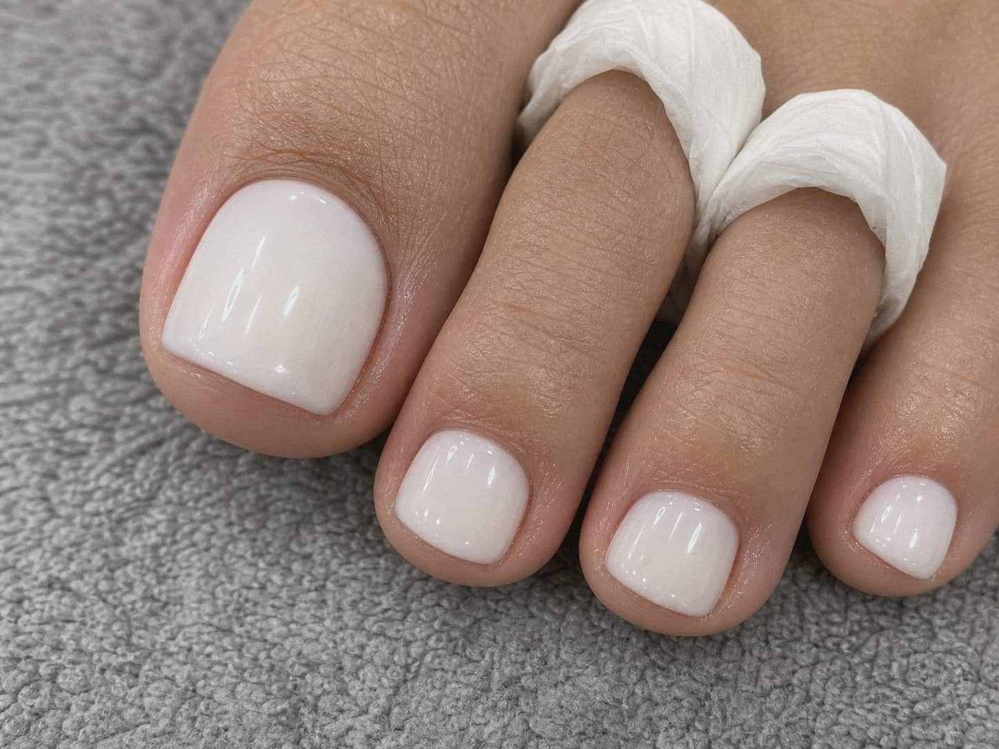 Tight close-up of a milky white gel pedicure with glossy short toenails and disposable toe separators.