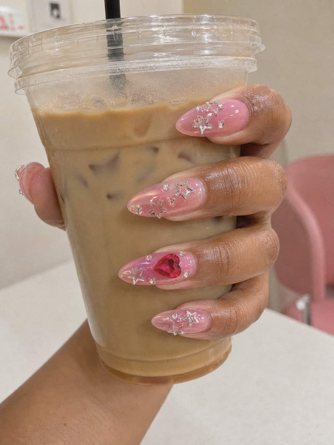 Freshly finished long almond pink ombre nails with silver stars and a red heart charm holding an iced coffee cup in an iPhone-style close-up.