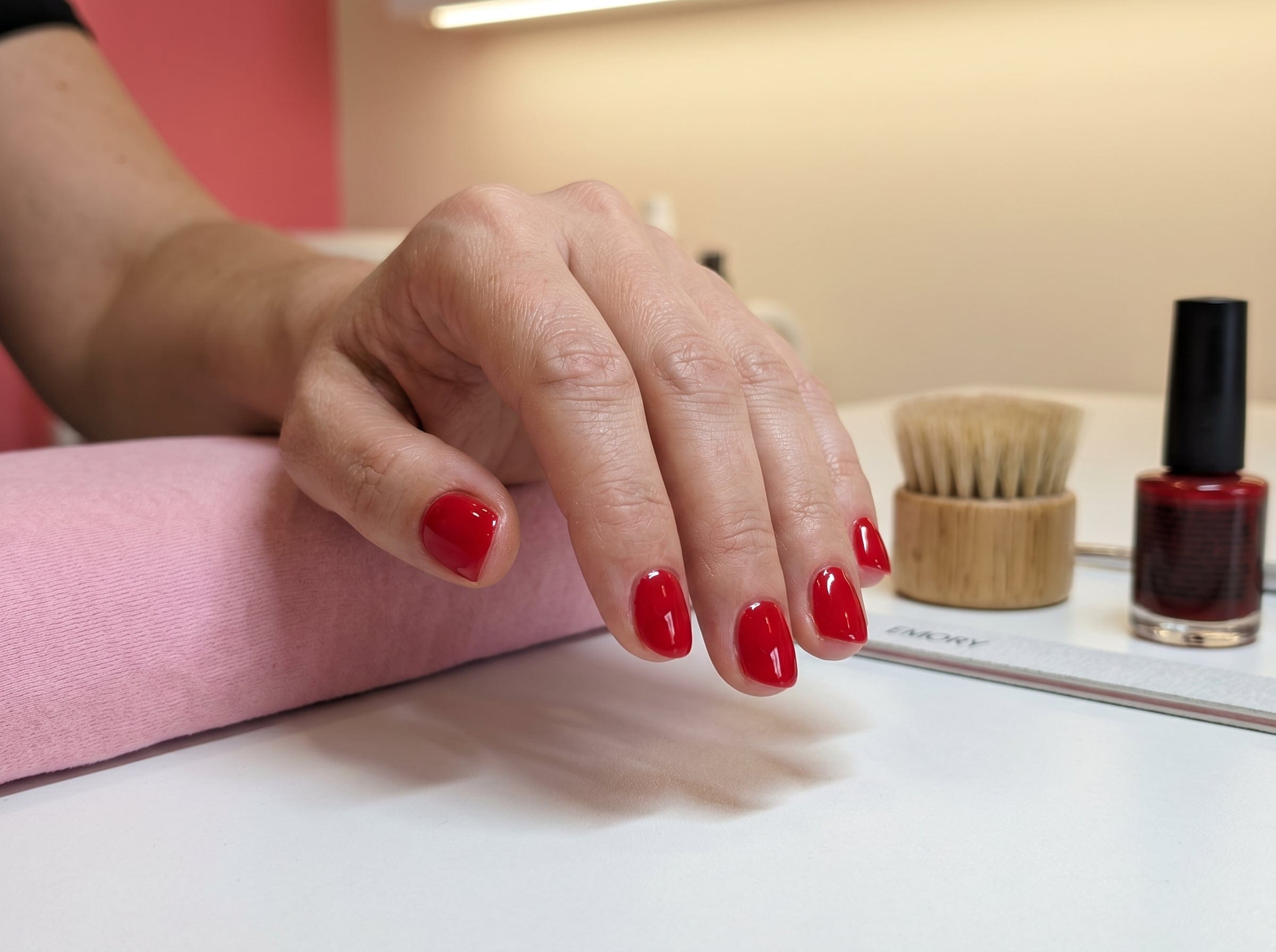 Glossy classic red gel manicure on short rounded nails resting on a pink cushion with a red polish bottle nearby.