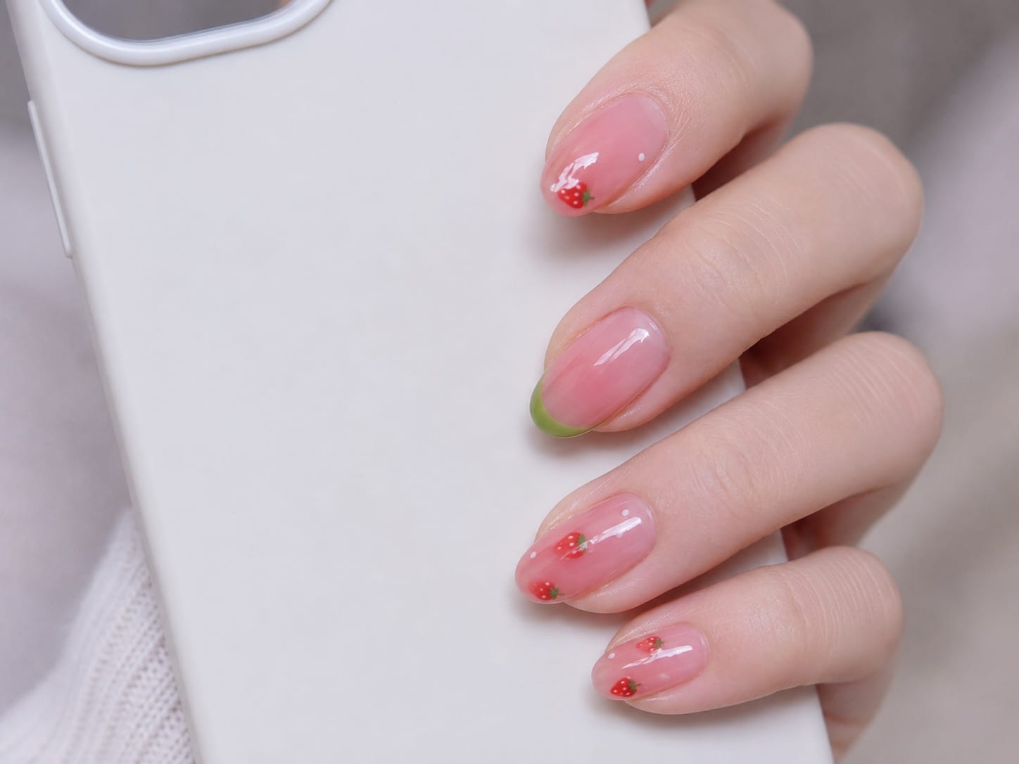 Short almond pink jelly nails with red strawberry art and a green French tip beside a white phone case.