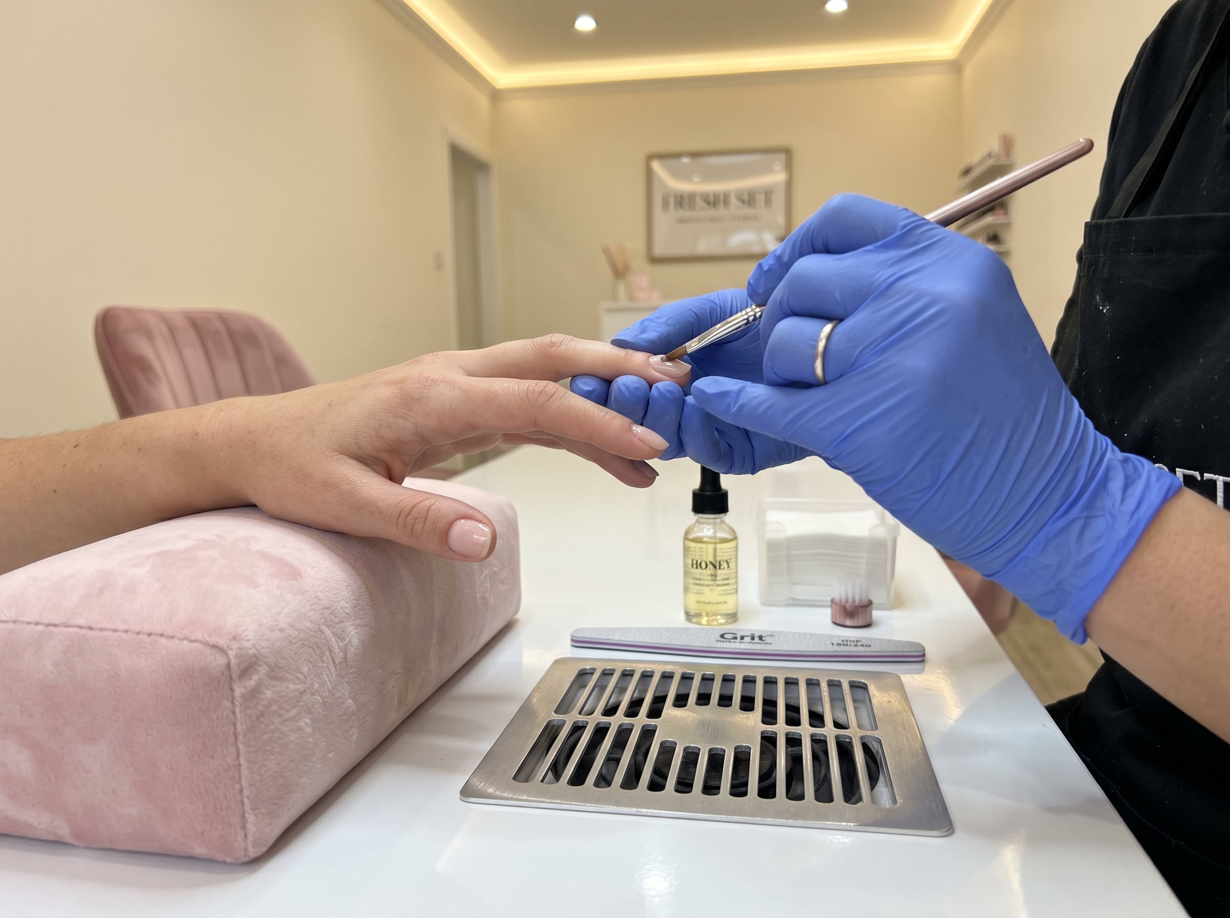 Tech in blue gloves applying BIAB builder gel at a salon table with a Fresh Set sign and oil bottle.