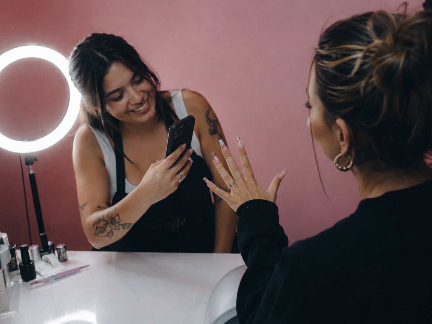 Smiling tech photographing a client's new manicure with a phone at the pink-wall ring-light vanity station.
